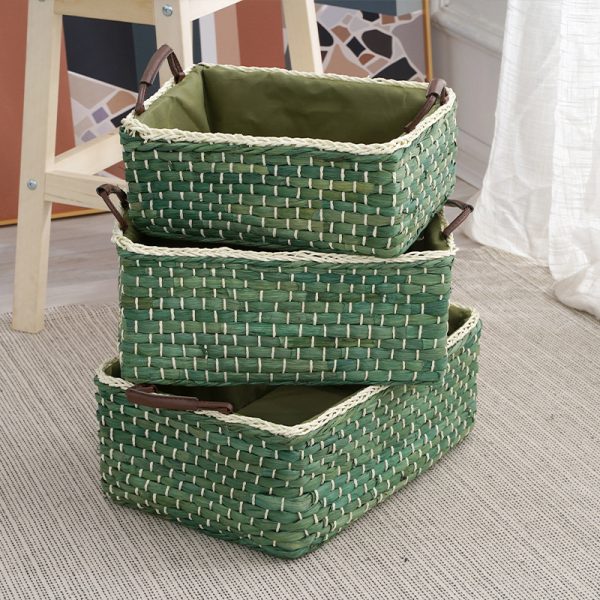 Three handwoven corn husk storage baskets in green, brown, and white, featuring leather handles and fabric liners, stacked on a textured rug in a modern living room setting.