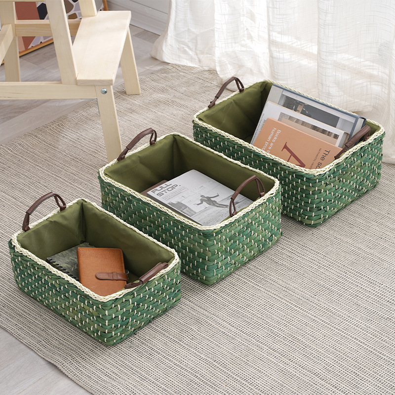 Three handwoven corn husk storage baskets in green, brown, and white, featuring leather handles and fabric liners, stacked on a textured rug in a modern living room setting.